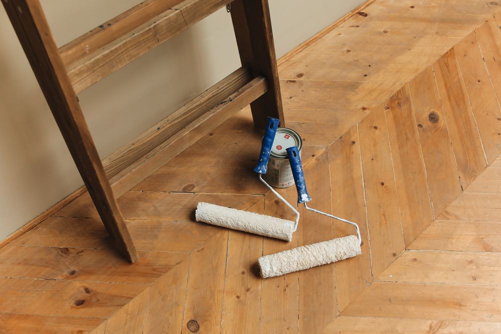 Paint rollers and a paint can on a wood floor beside a ladder